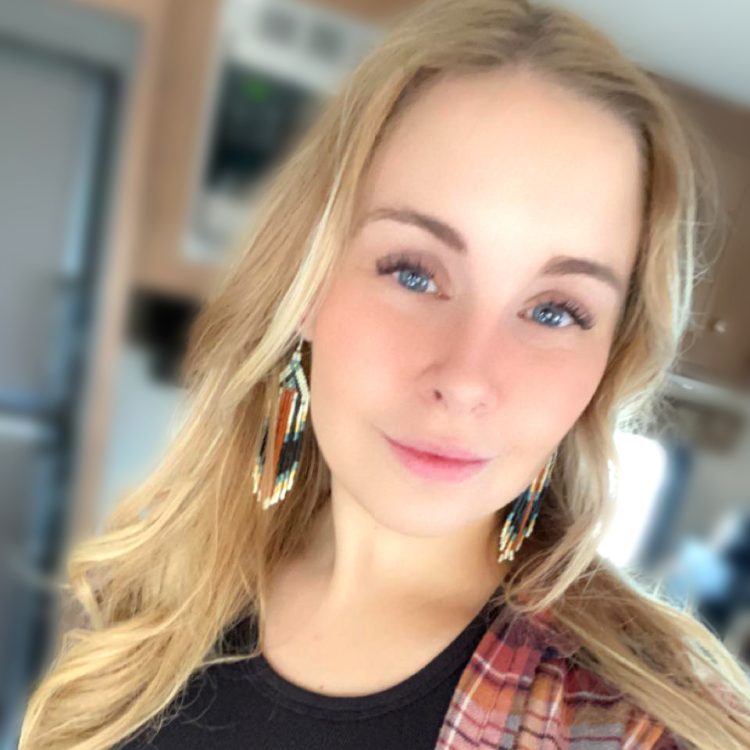 A young woman with long blonde hair wears dangling beaded earrings and a plaid shirt, smiling slightly while looking at the camera indoors.