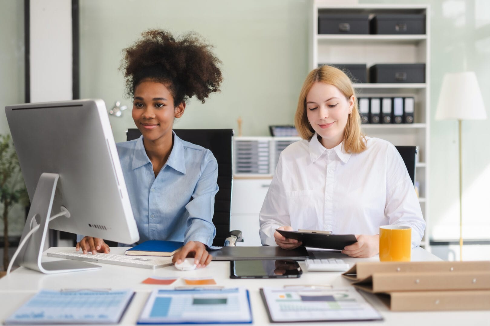 Two professionally dressed, diverse females sit at a desk. Spreadsheets and binders can be seen on the desk.
