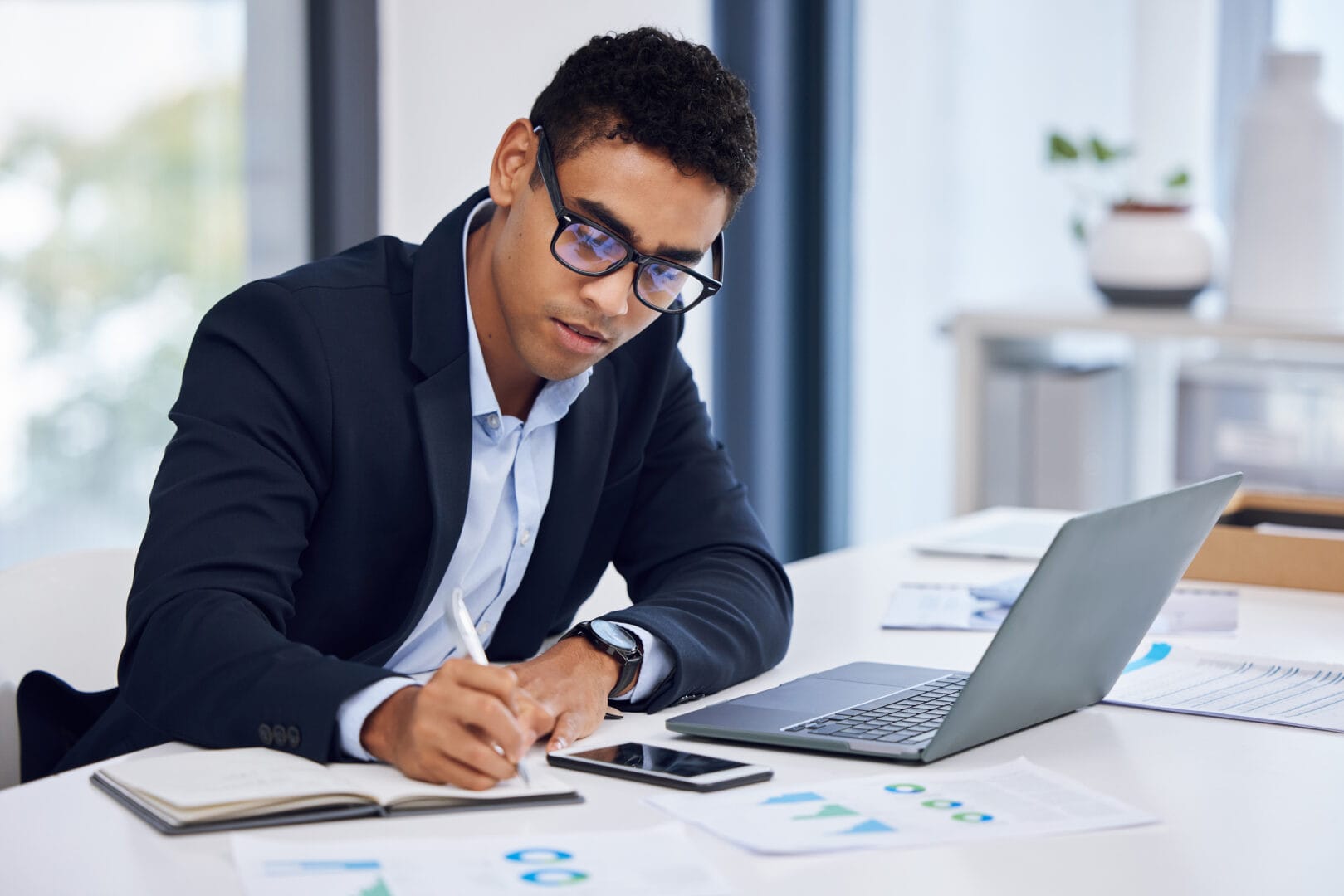 A young, professionally dressed male sits at a desk where a laptop, spreadsheets, and a smartphone can be seen.